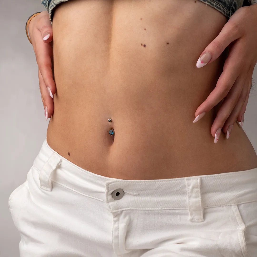 Detail of the Orb 7mm London Blue Topaz belly button ring in a navel piercing, framed by a grey crop top and white jeans